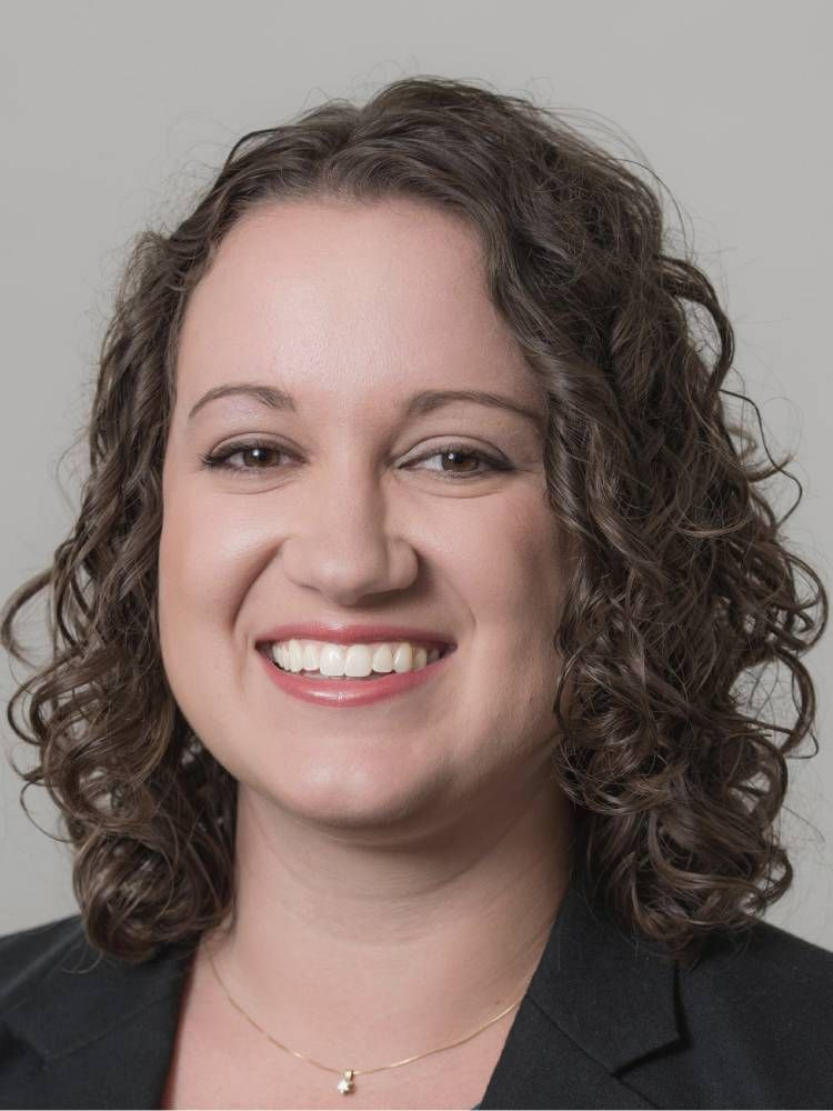 Woman with curly brown hair smiles. Wearing a dark suit jacket and a teal undershirt, a gold necklace.