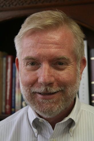 Smiling, middle-aged man with blonde hair and beard, wearing a light-colored button-down shirt; bookshelves in the background.