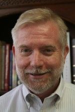 Smiling, middle-aged man with blonde hair and beard, wearing a light-colored button-down shirt; bookshelves in the background.