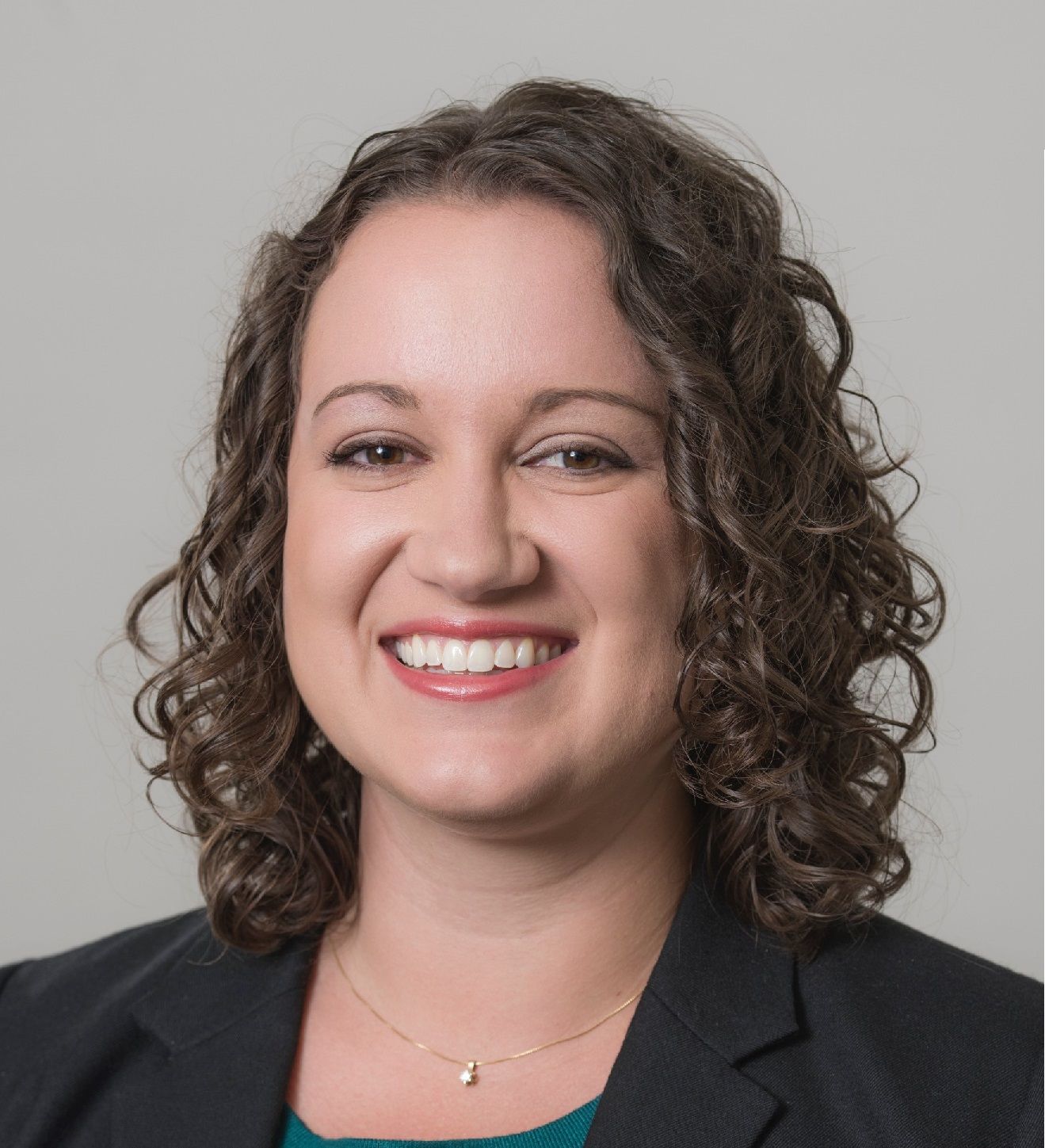 Woman with curly brown hair smiles. Wearing a dark suit jacket and a teal undershirt, a gold necklace.