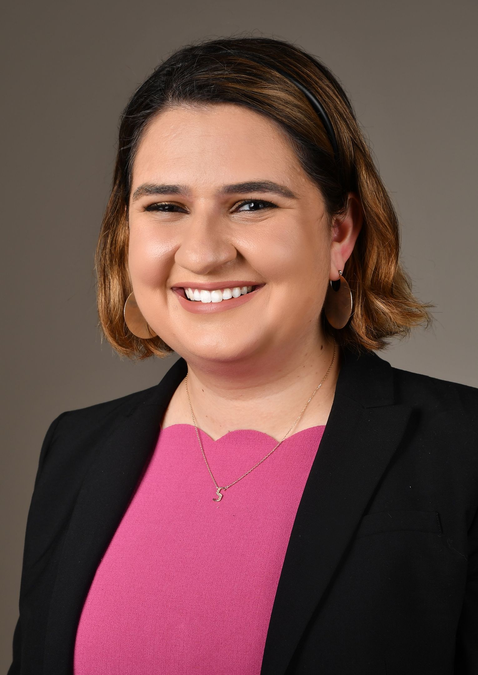 Woman with dark hair, smiling, wearing a pink shirt, black blazer, and gold jewelry, against a grey background.