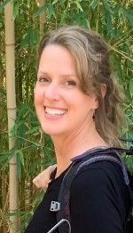 Woman smiles, outdoors. Wearing black shirt and backpack straps. Light skin, blonde hair. Bamboo background.