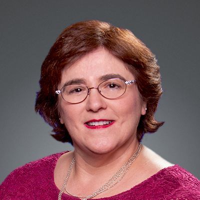 Woman with glasses, smiling, wearing a magenta sweater, necklace, set against a blue background.