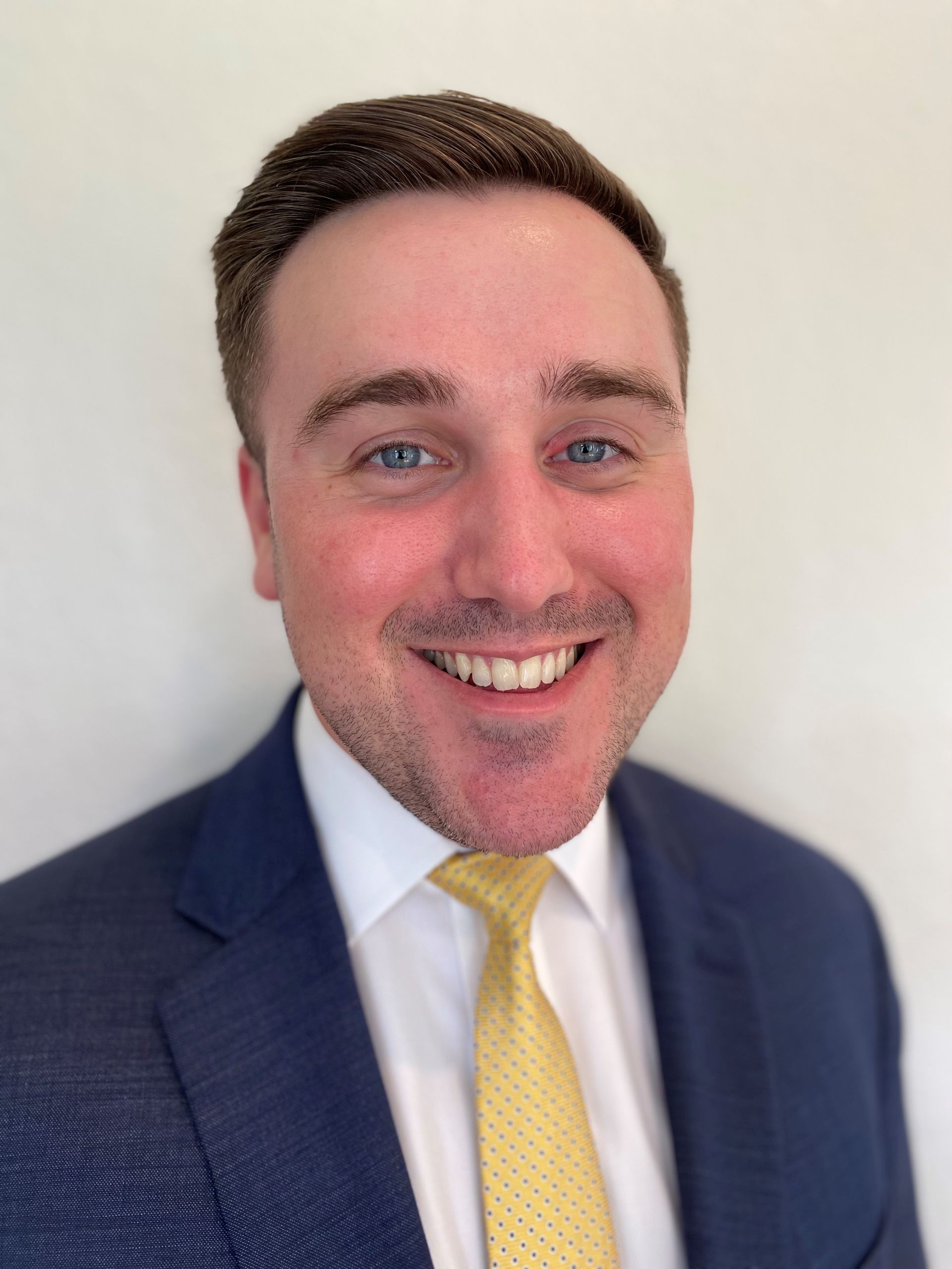 Man in navy suit, white shirt, and yellow tie smiles broadly, headshot.