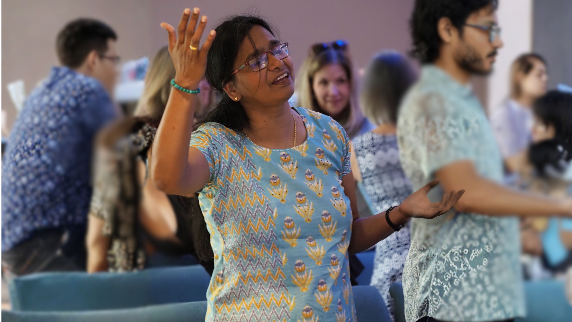 Woman in blue patterned shirt raising hand, inside a building with others.