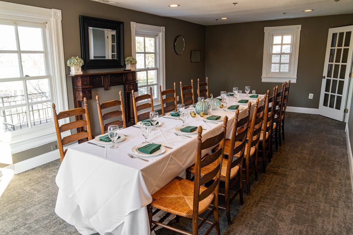 Long dining table set for a meal in a room with wood chairs, windows, and dark walls.