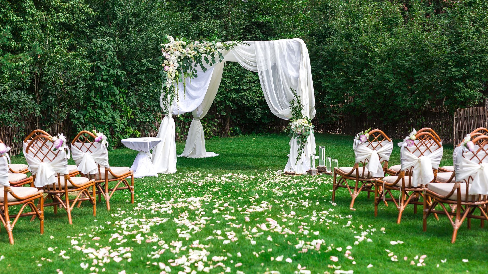 Wedding ceremony setup in a green yard with white arch, chairs, and floral petals on grass.