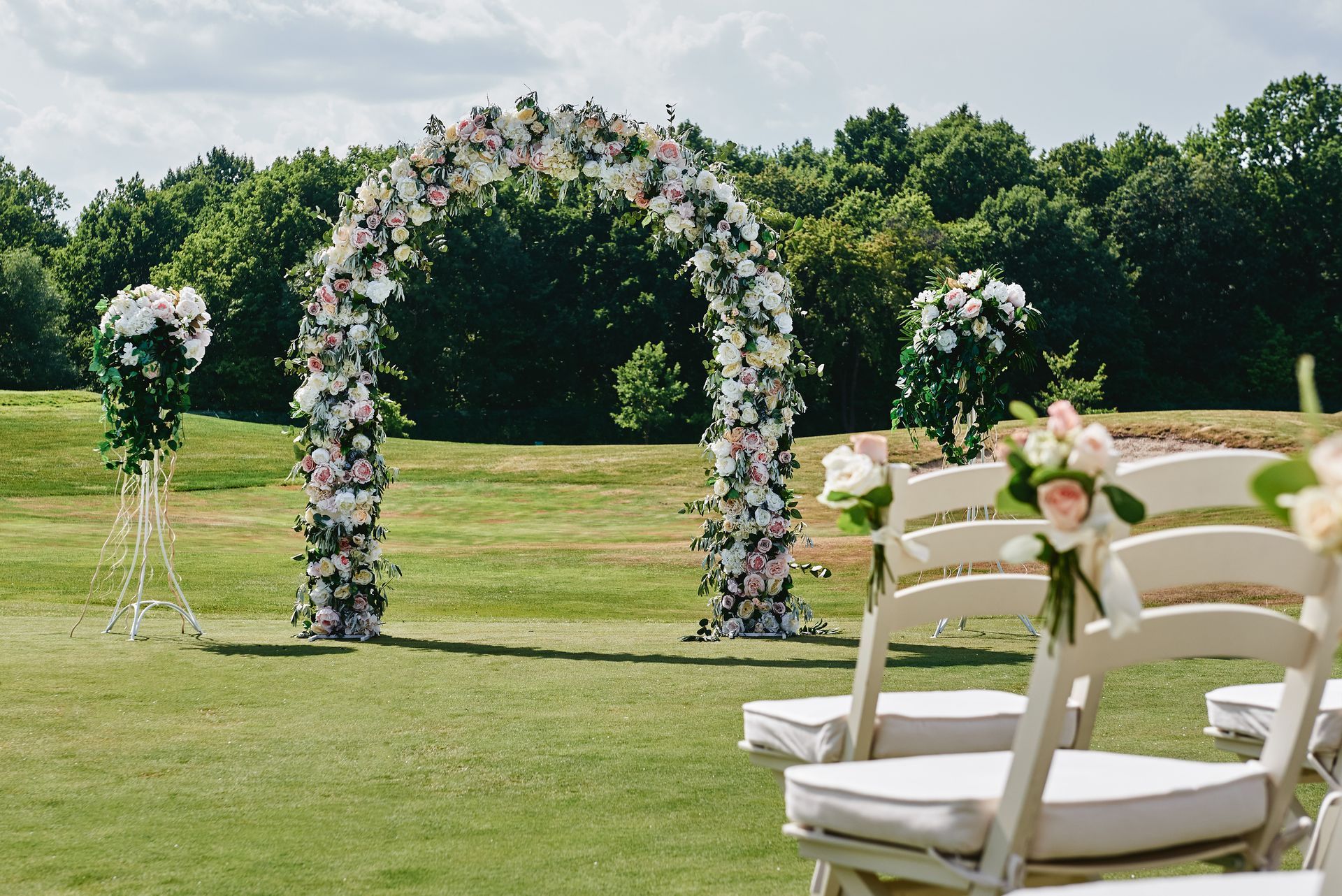 Wedding ceremony setup with floral arch and chairs on a grassy field, trees in background.