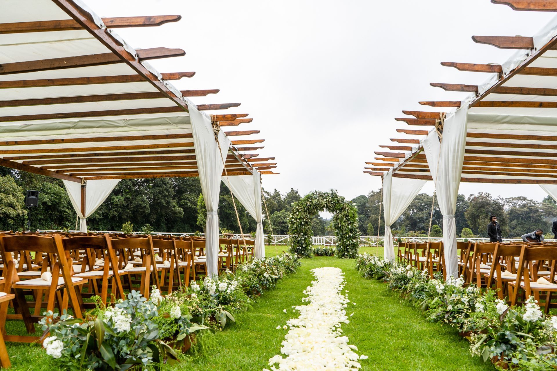 Outdoor wedding ceremony setup with rows of chairs, floral aisle, and draped pergolas.