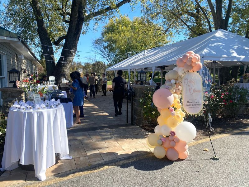 Outdoor event with balloons, tables, and people near a white tent.