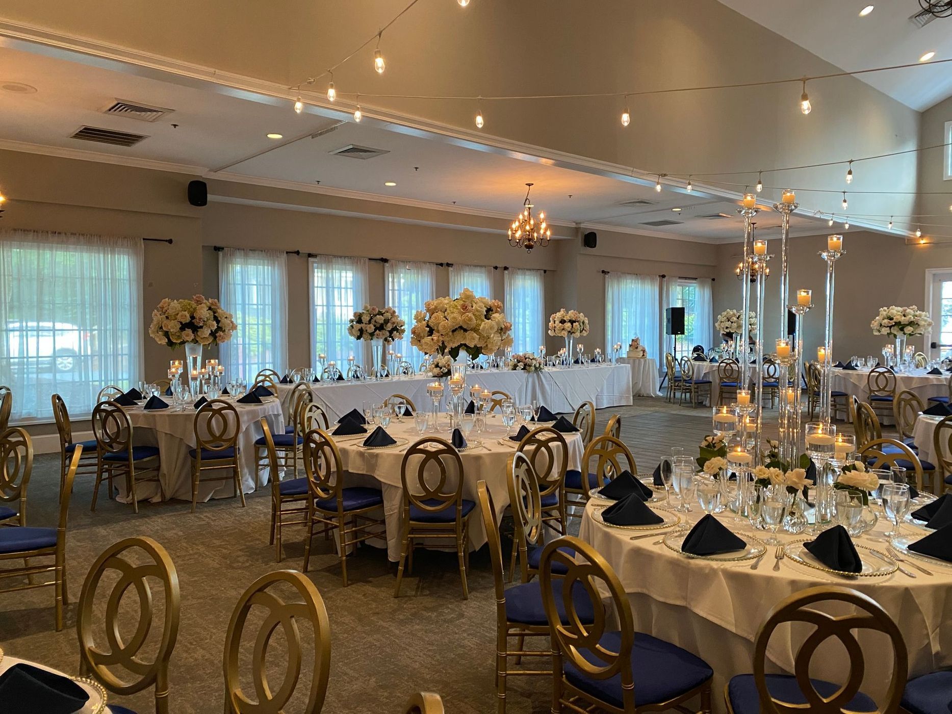 Elegant banquet hall with round tables set for a formal event. Tables have white linens, blue chairs, and tall floral centerpieces.