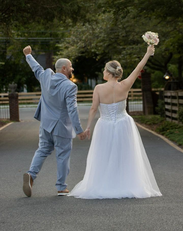 Newlyweds raise arms, walking away down a paved path; the bride holds a bouquet.