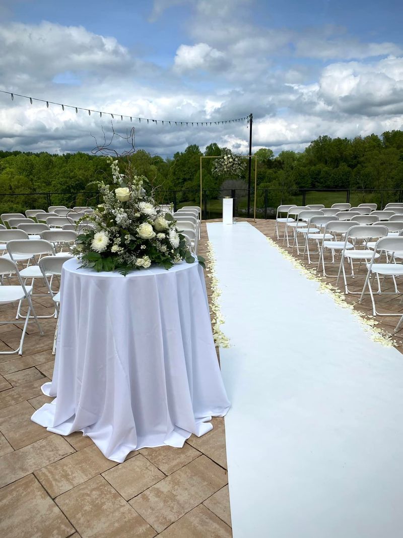 Outdoor wedding ceremony setup with white aisle runner, flower arrangement on draped table, and rows of white chairs.