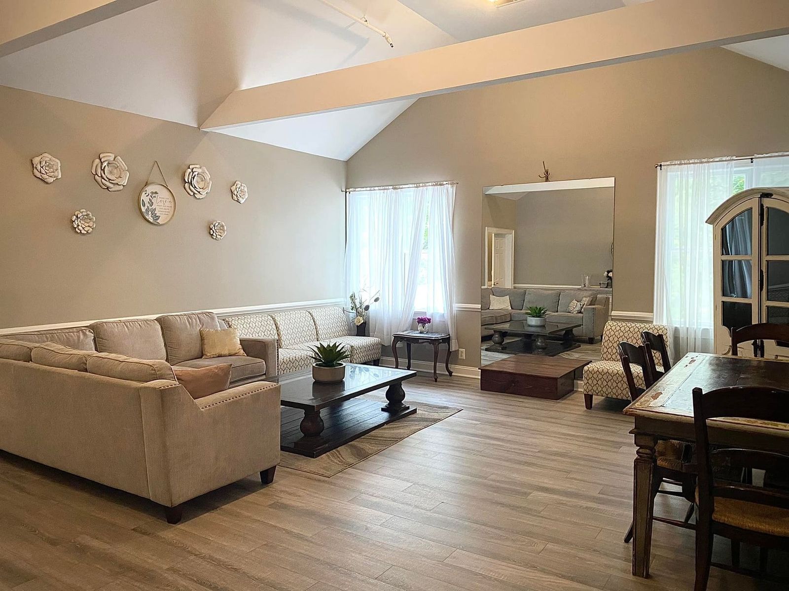 Living room with a large sofa, tall mirror, and dining table, neutral colors and hardwood floors.