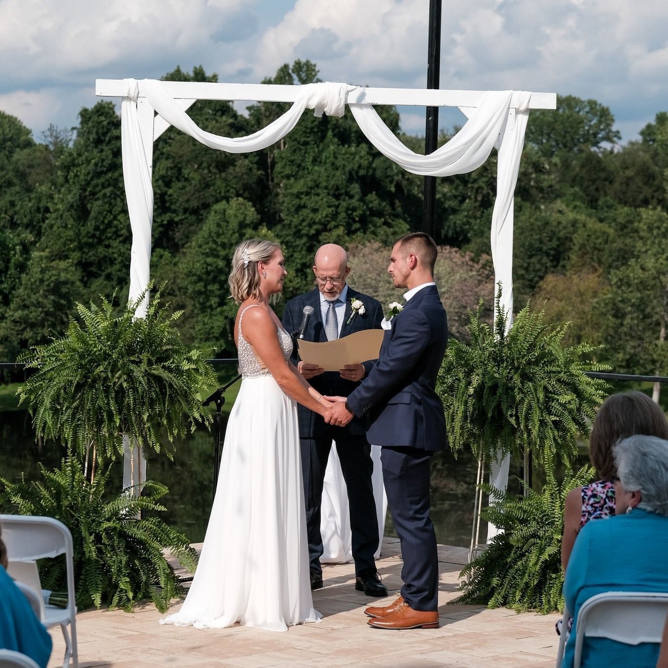Bride and groom holding hands during outdoor wedding ceremony; officiant reads.