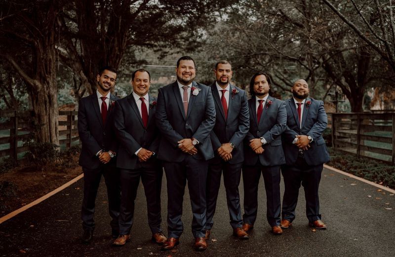 Six men in navy suits and red ties stand on a paved path, smiling.