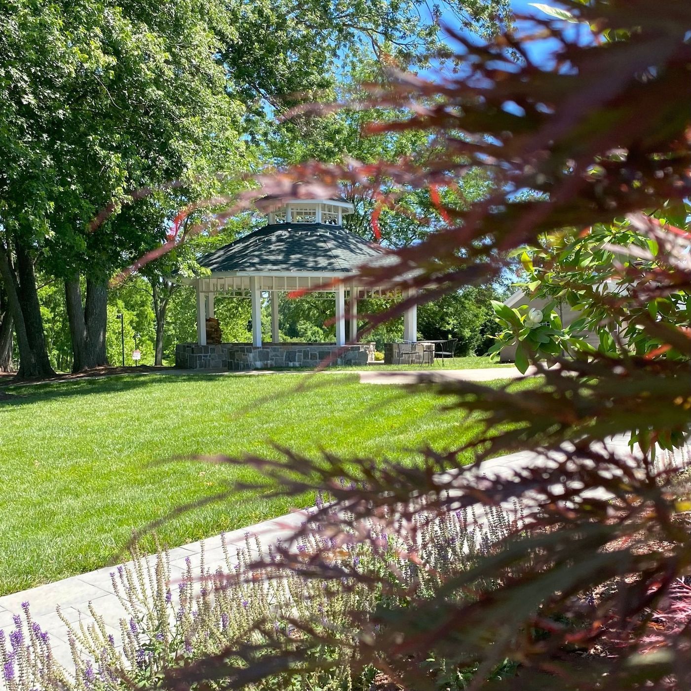 Gazebo in a sunny park with a stone base, green grass, and trees. Red leaves in the foreground.