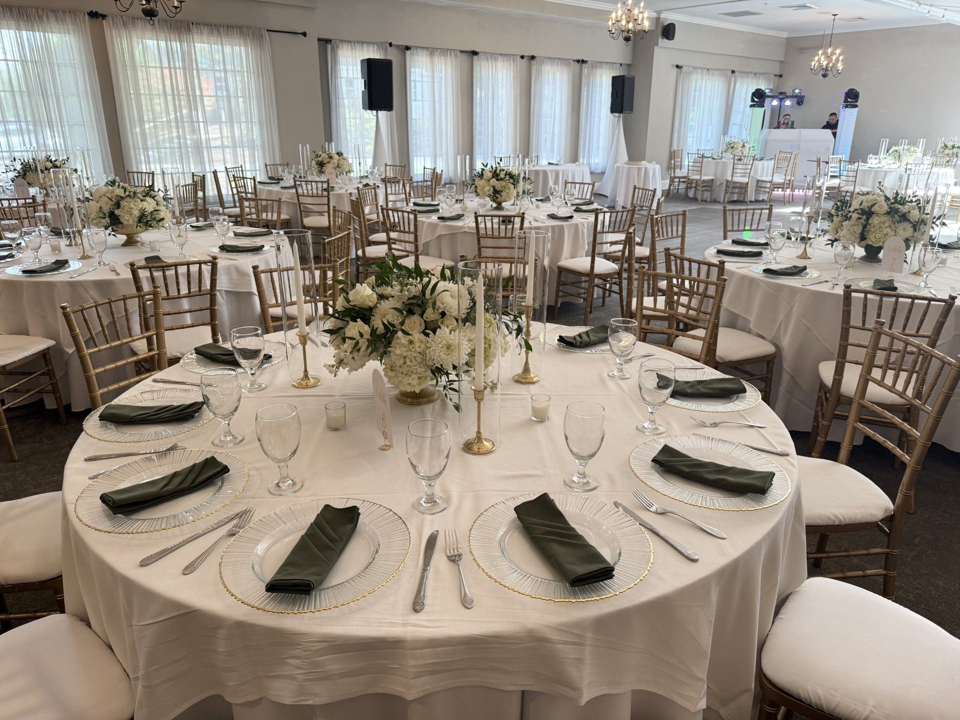 People mingle at a well-lit event in a large room with chandeliers, tables, and windows.
