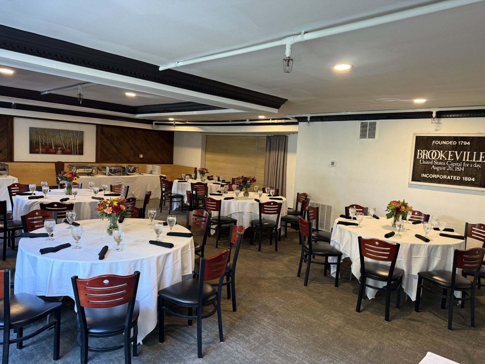 Restaurant dining room with round tables set for a meal. Fireplace, windows, and chairs are visible.