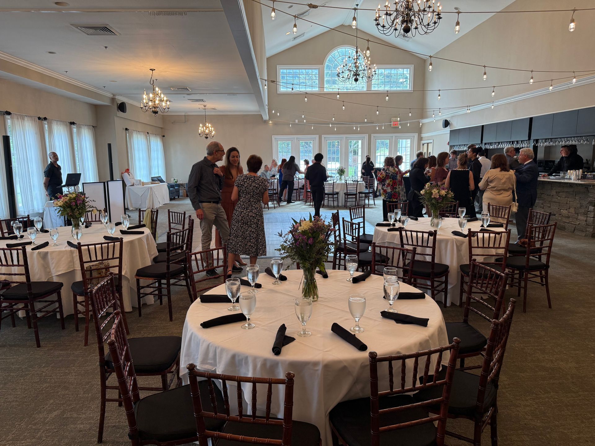 Wedding reception hall with round tables set for guests; people mingle, chatter; chandeliers hang.