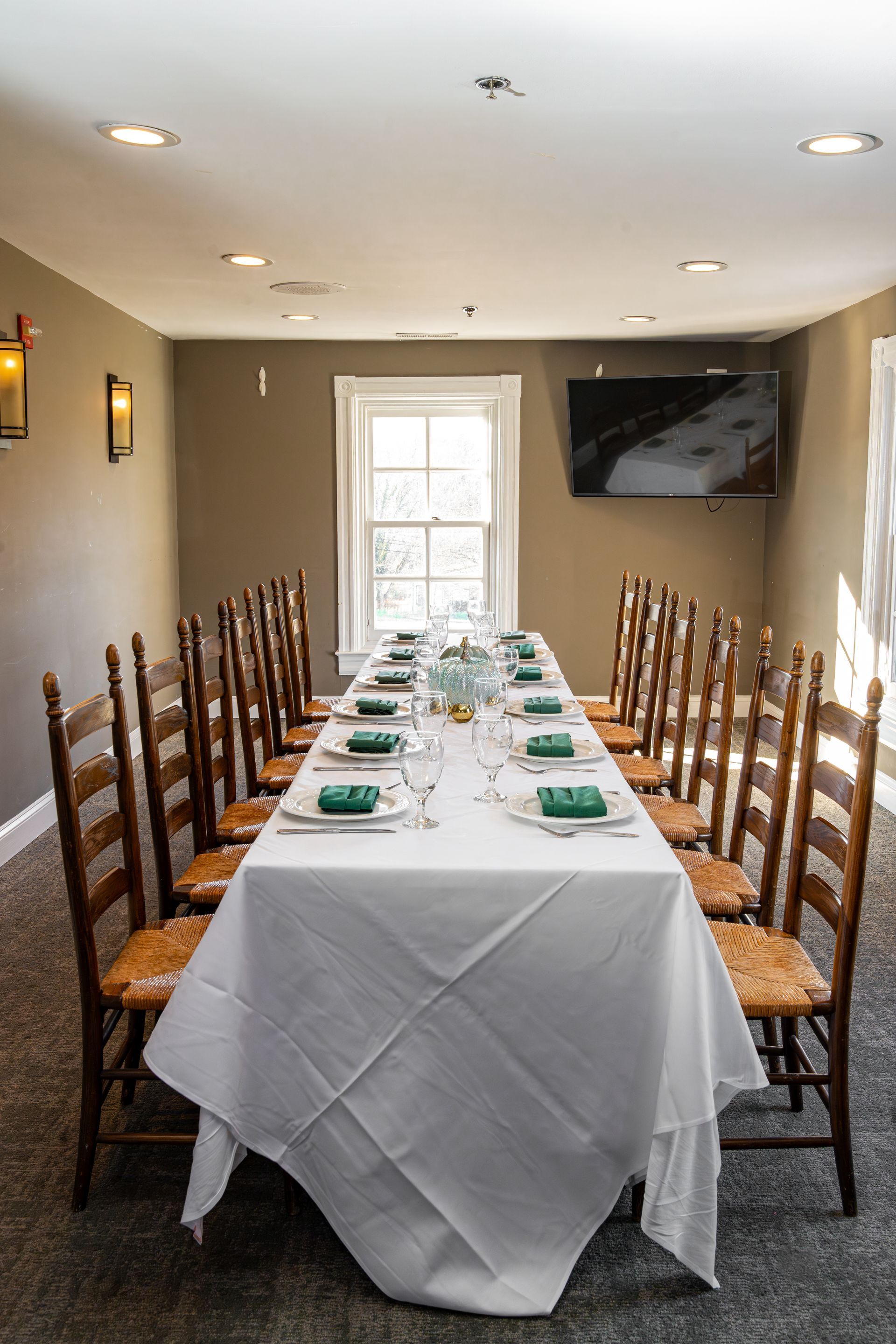 Long dining table set for a meal in a room with chairs, a window, and a TV.