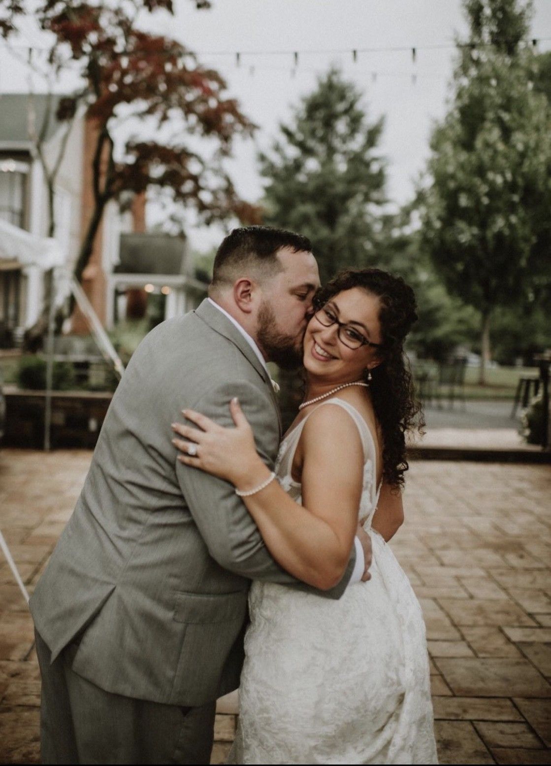 Groom kisses bride's cheek, embracing her, outdoors. She smiles, wearing glasses and a wedding dress.