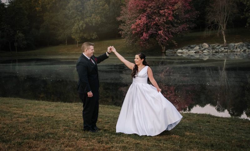Newlyweds dancing by a lake, the bride in a white gown and the groom in a suit, under a tree.