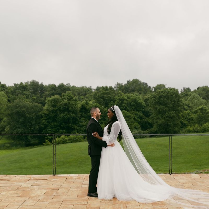 Couple in formal wear, embracing on a patio overlooking a green landscape. Overcast sky.