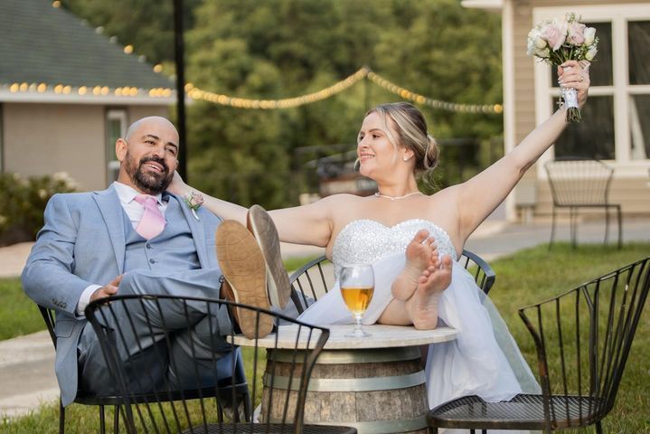 Newlyweds relaxing, feet up on table, holding bouquet and beer. Outdoors at wedding venue.