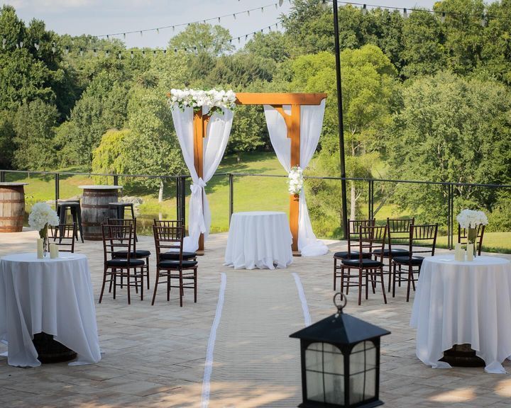 Outdoor wedding ceremony setup with white linens, wooden arbor, and surrounding trees.