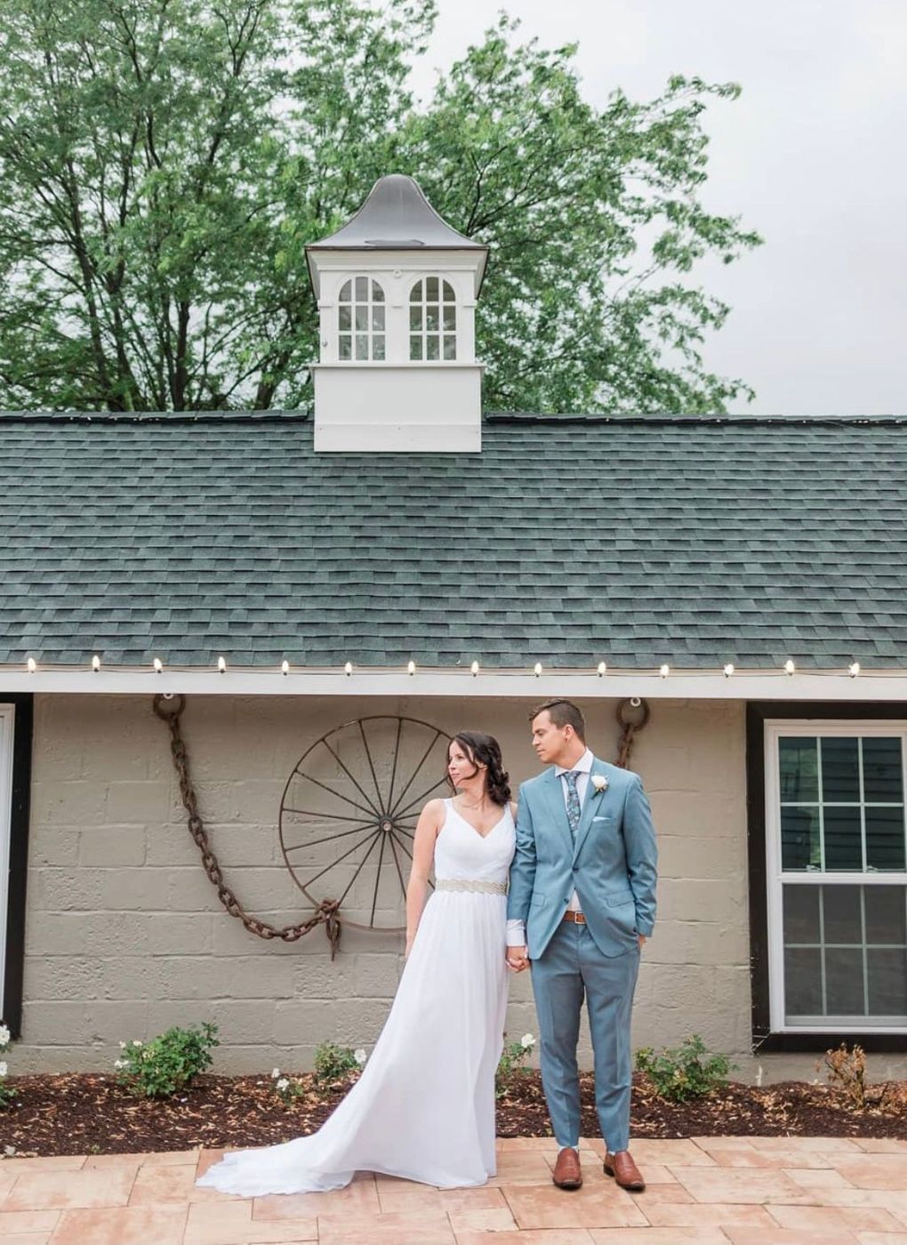 Bride and groom hold hands in front of a barn, she in a white gown, he in a blue suit.