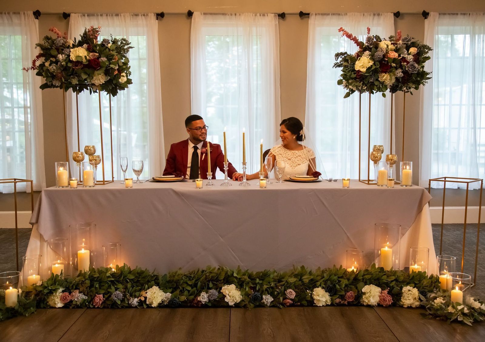 Newlyweds seated at a table decorated with flowers and candles; both are holding hands.