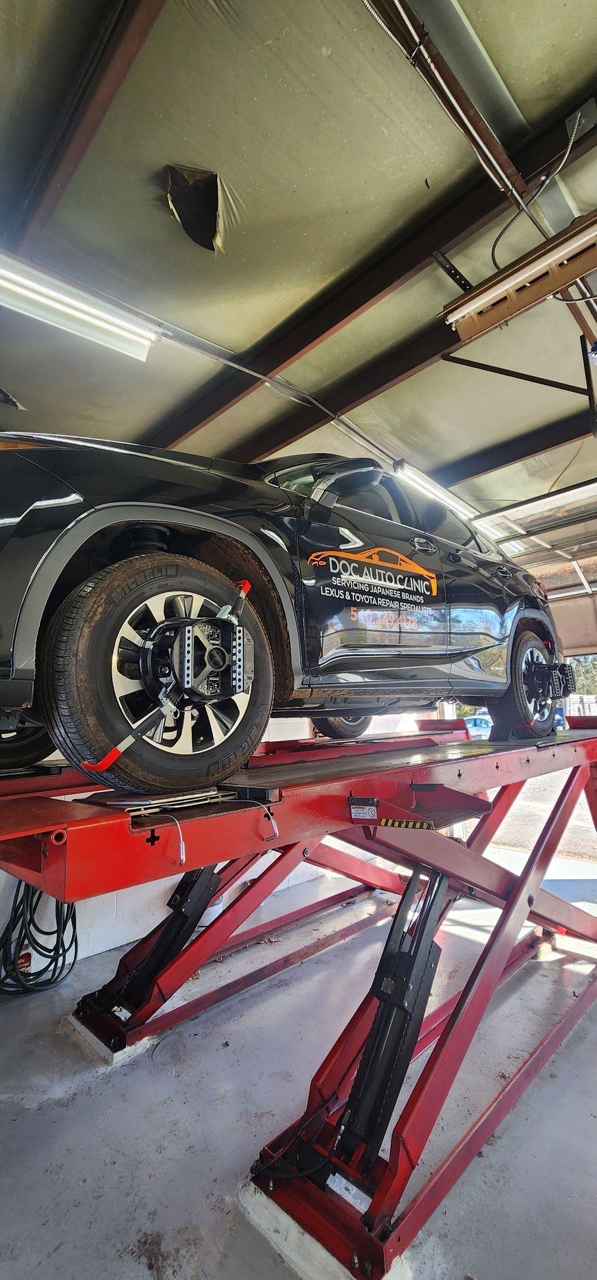 A black car on a red scissor lift inside a garage, undergoing wheel alignment. | Ish Auto Clinic