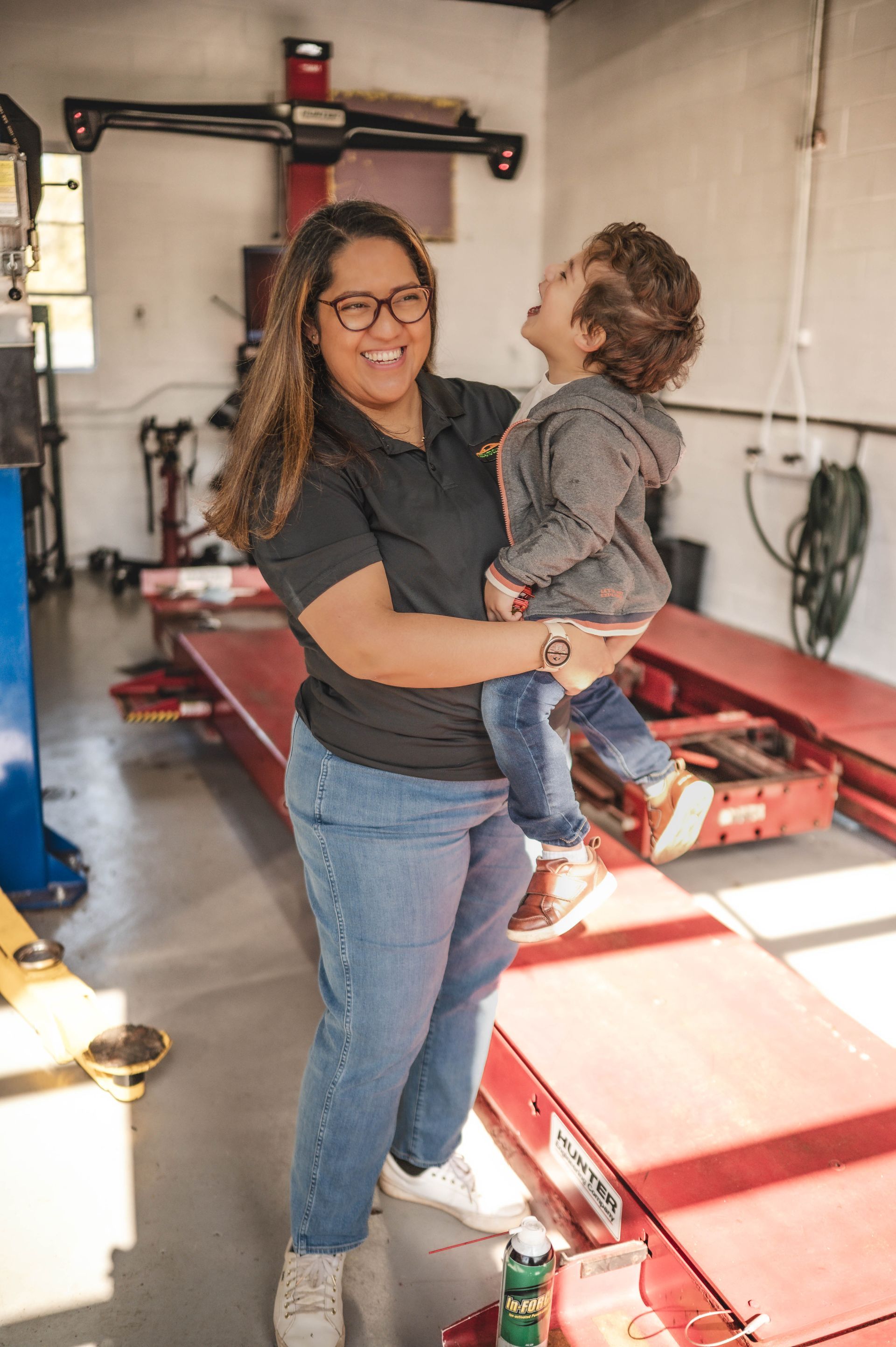 Woman holding a laughing toddler in a car repair shop; she smiles. | Ish Auto Clinic