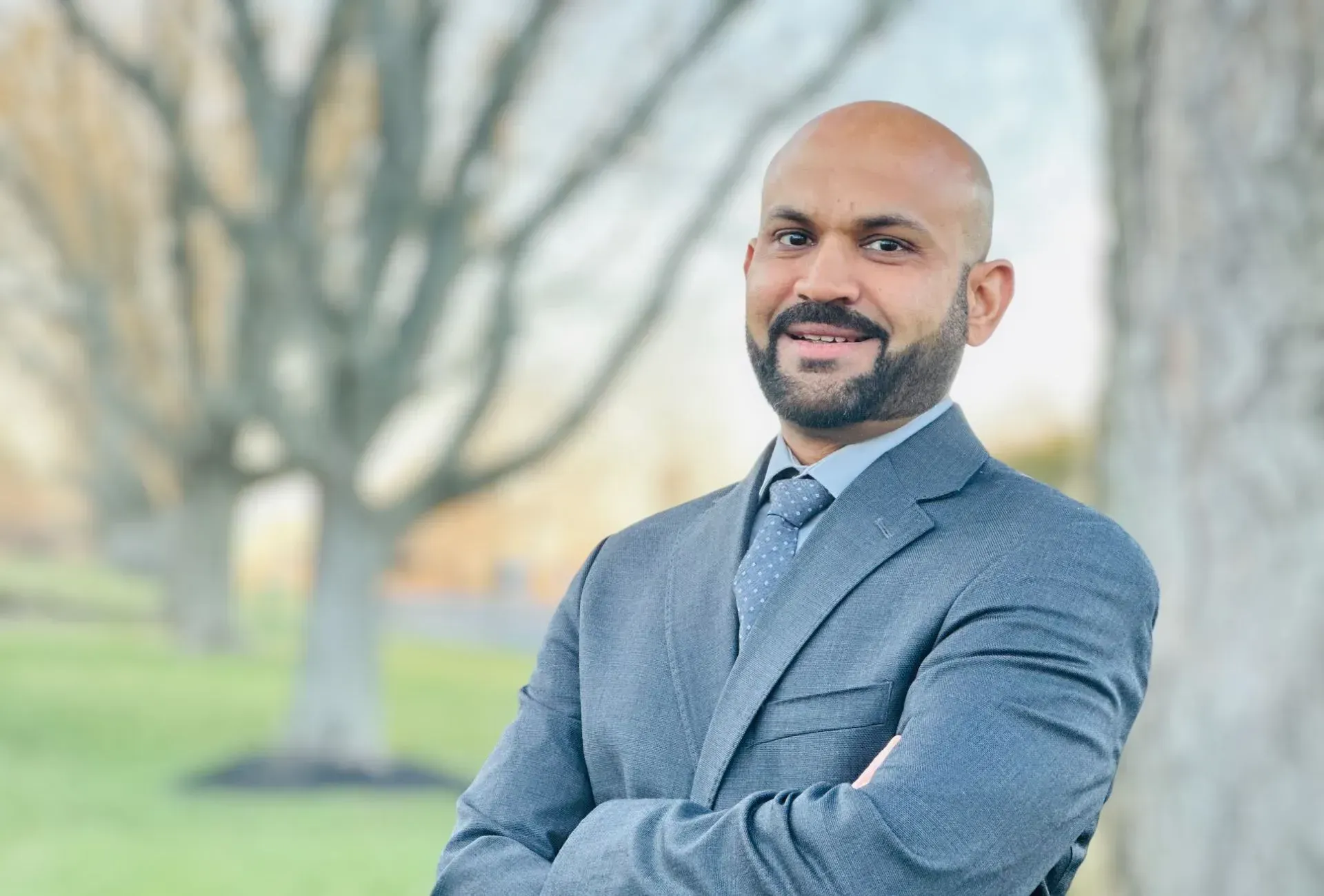 A man in a suit and tie is standing in front of a tree with his arms crossed.