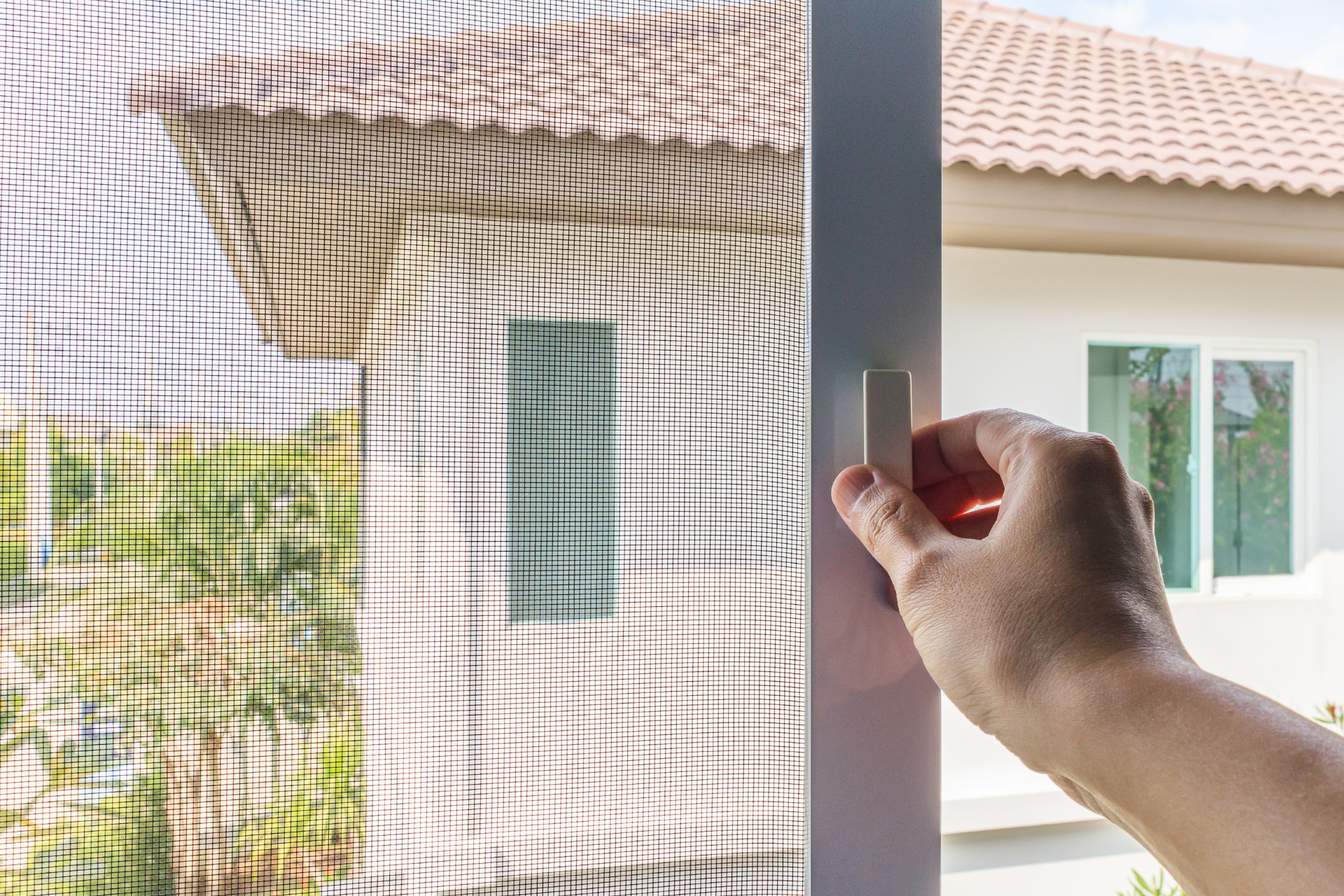 A person's hand opening a screen door latch, blurred background of a house and yard.