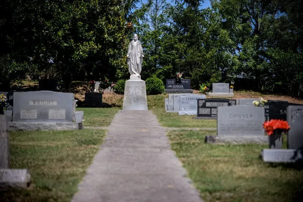 Stone statue atop a pedestal, centered on a walkway in a cemetery, with headstones and trees.