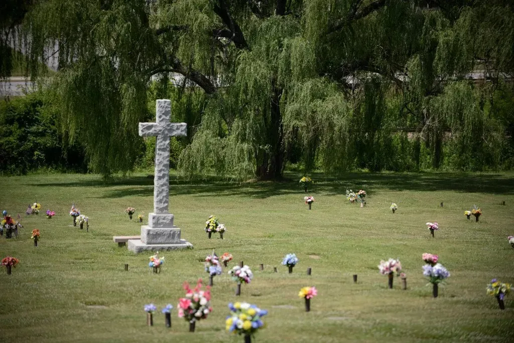 Cross-shaped monument in a green cemetery field, weeping willow tree in the background, colorful flower arrangements.
