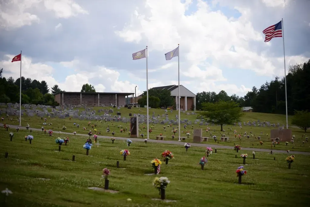 Cemetery with headstones and flags under a cloudy sky. Memorial building in background.