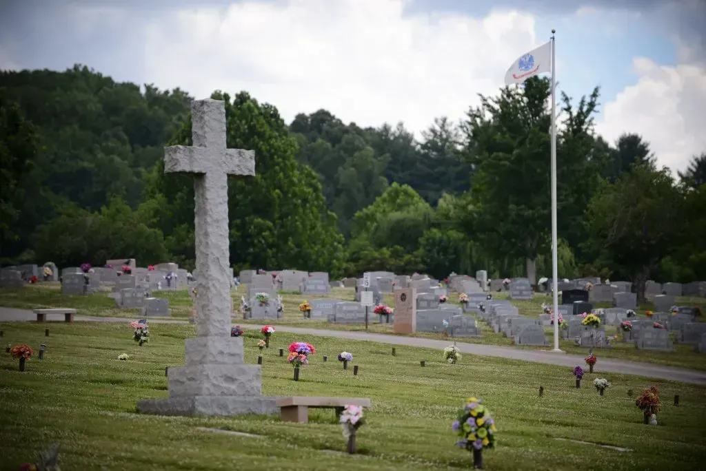Stone cross in a cemetery, surrounded by headstones and flowers. A flag waves in the background.