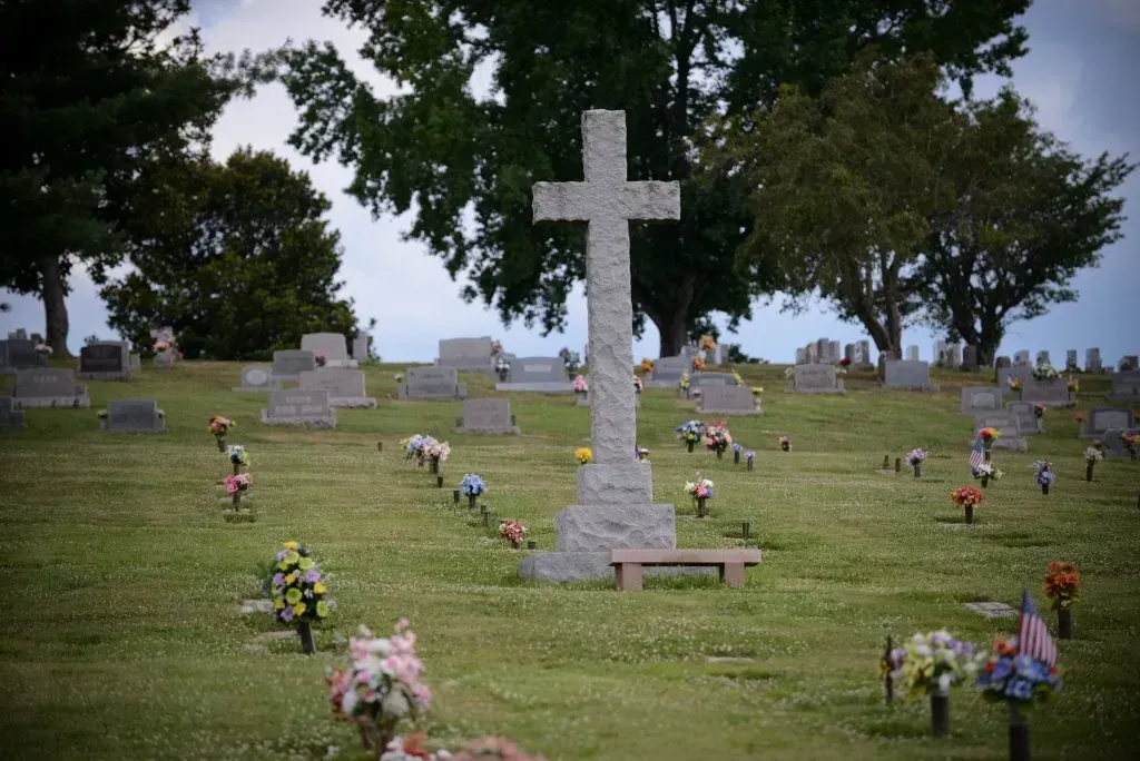 Stone cross in a grassy cemetery, surrounded by tombstones and trees.