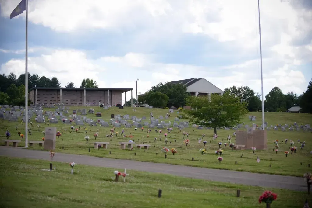 Cemetery with numerous headstones on a grassy hill; building and flags in the background; sunny day.