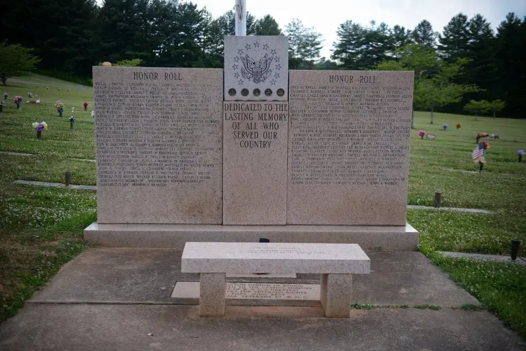 War memorial with names, a coat of arms, and a stone bench in a cemetery setting.