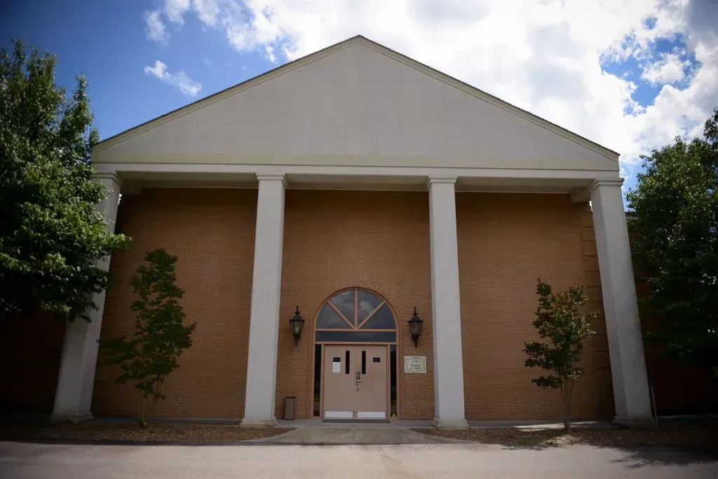 Brick building with a white triangular roof and three large pillars. Two trees flank the entrance.