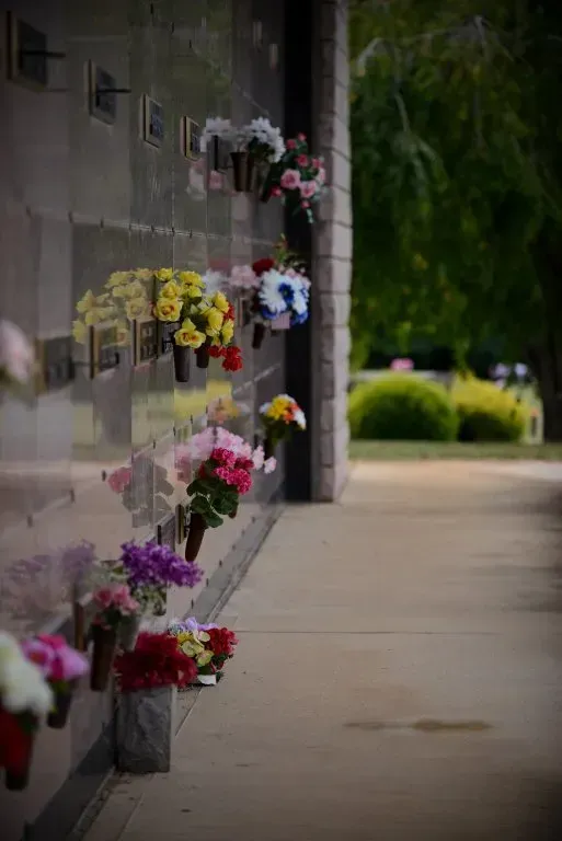 Cemetery columbarium with colorful flower arrangements in front of niches. Green trees and foliage are in the background.