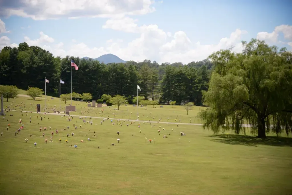 Green cemetery with headstones and flags on a sunny day; mountains in the background.
