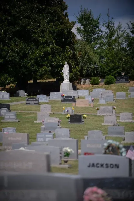 Cemetery with many headstones on a grassy hill; a white statue is prominent in the distance.
