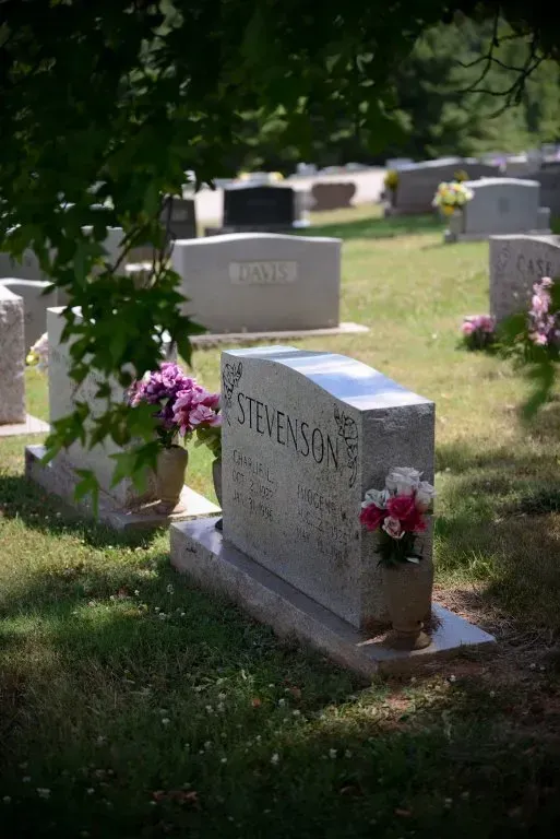 Gravestone in a cemetery, surrounded by other headstones and green grass; flowers on the grave.