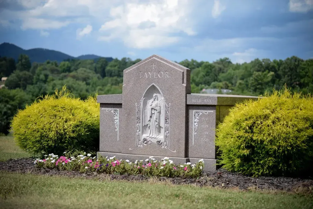 Headstone in a cemetery with a statue of an angel, surrounded by greenery and mountains.
