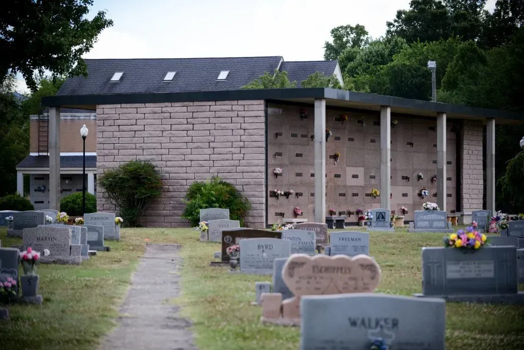 Cemetery with a mausoleum in the background and headstones in the foreground. Green grass and trees surround.